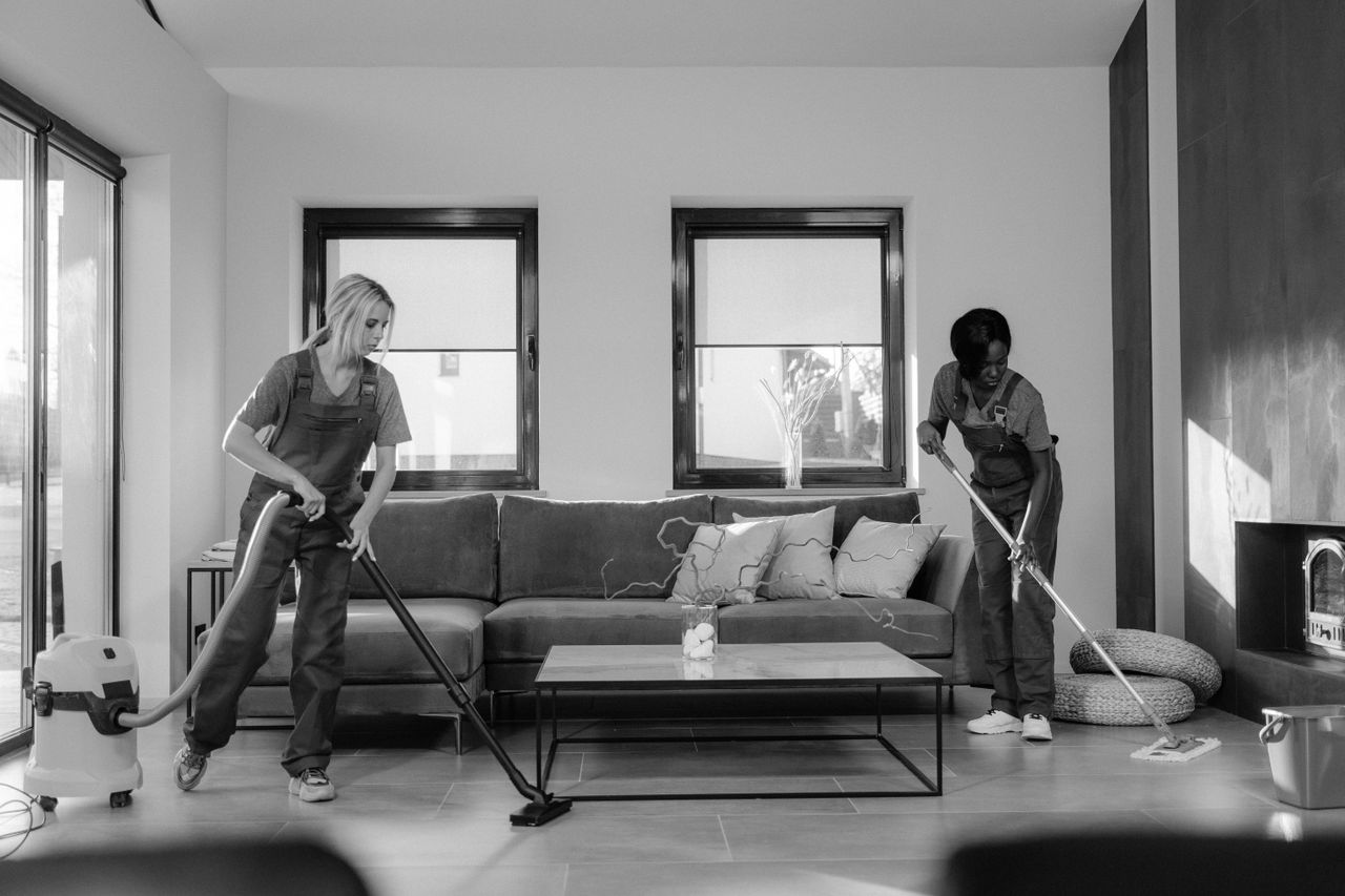 heros-img Two women in work clothes are cleaning a modern living room with a vacuum and a mop.